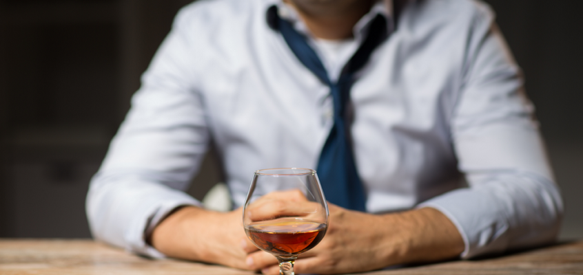 A man sits at a wooden table with a snifter of amber liquor in the foreground.