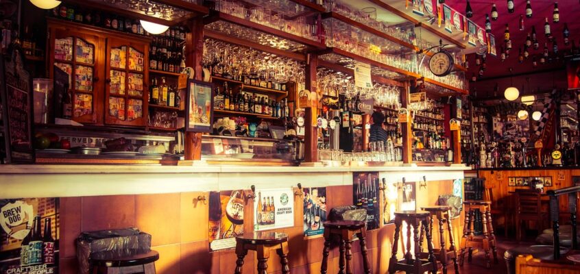 A detailed view of a crowded bar with wooden stools, hanging beer bottles, and glassware.