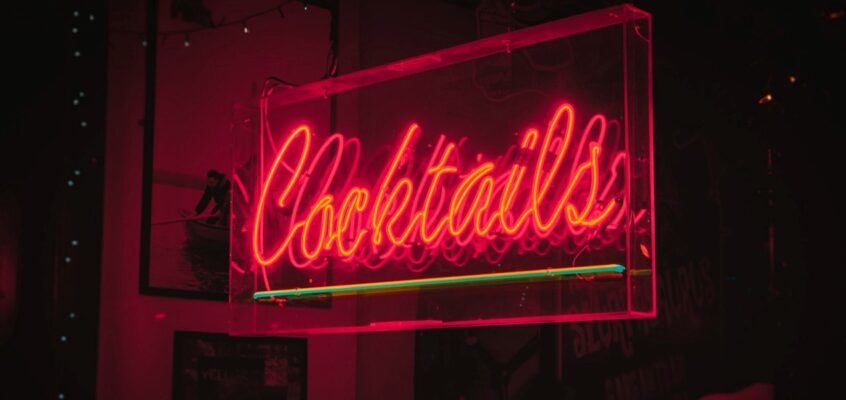 A glowing pink and red neon sign that reads "Cocktails" encased in a clear acrylic box within a dimly lit bar.