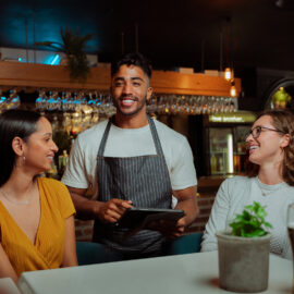 A male bartender in a striped apron smiles while using a tablet to assist two female patrons at a bar.