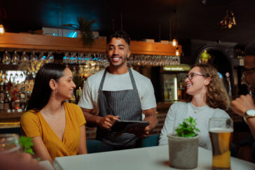 A male bartender in a striped apron smiles while using a tablet to assist two female patrons at a bar.