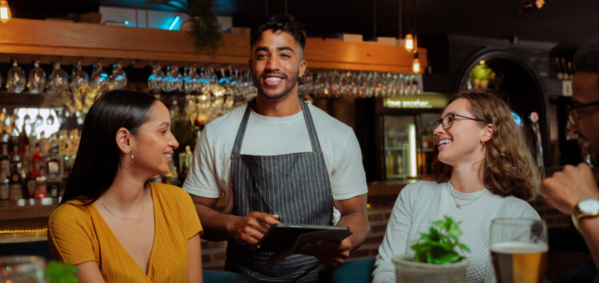A male bartender in a striped apron smiles while using a tablet to assist two female patrons at a bar.
