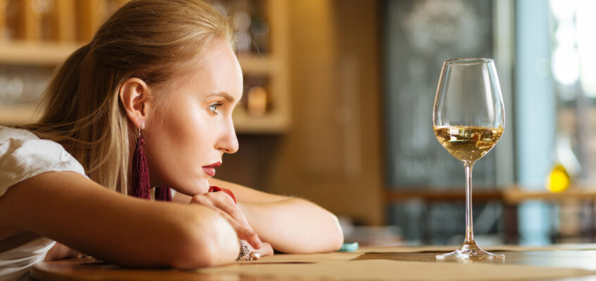 A blonde woman leaning her head on her arms on a bar table next to a glass of white wine.
