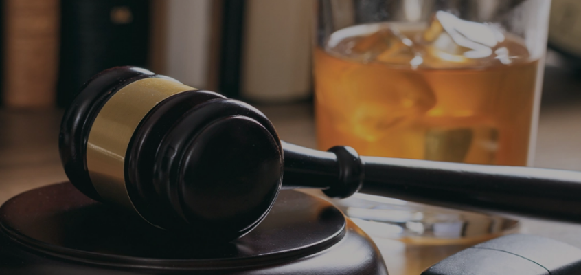 A judge's gavel and car keys on a desk with a glass of amber liquor in the background.