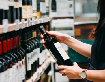 A customer or employee in a retail store holding a bottle of wine while browsing a well-stocked shelf of red wine.
