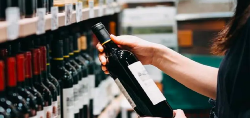 A customer or employee in a retail store holding a bottle of wine while browsing a well-stocked shelf of red wine.